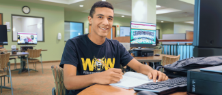 Boy sitting at computer taking notes in a notebook, smiling at the camera